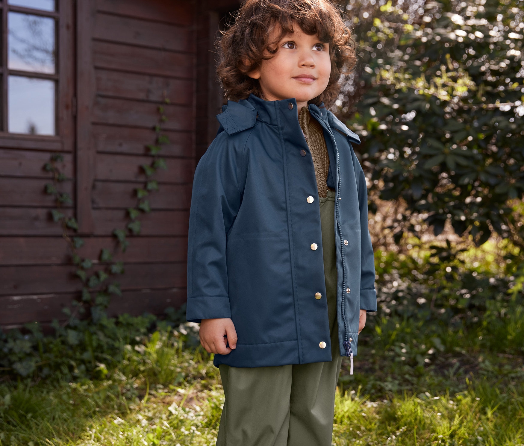 Egy fiú áll kint a fűben, gyerek gumicsizmát, kisgyerek esőnadrágot és kisgyerek esőkabátot visel.