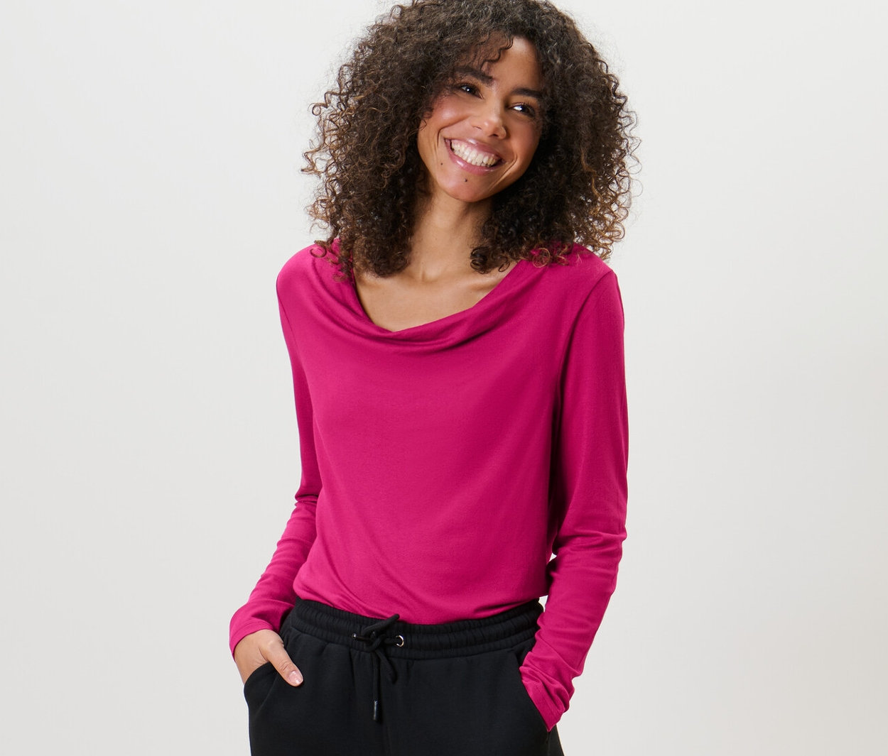 Göndör hajú nő málnapiros női vízesés nyakú pólóblúzt és fekete nadrágot visel.