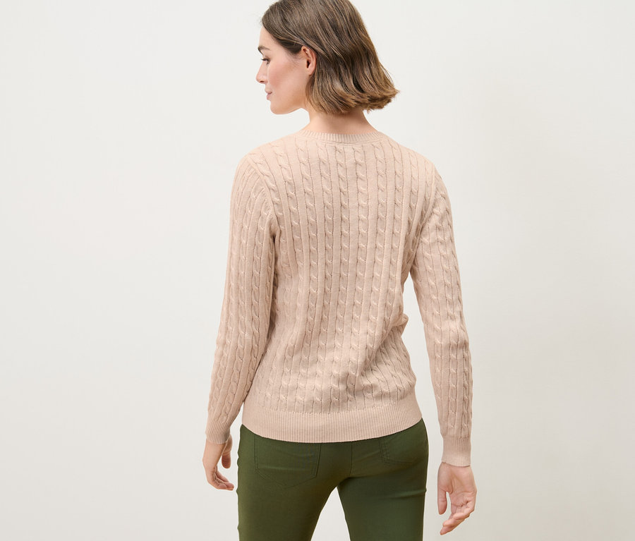 Barna hajú nő pózol háttal a kamerának, krémszínű, fonott mintás kötött pulóvert visel.