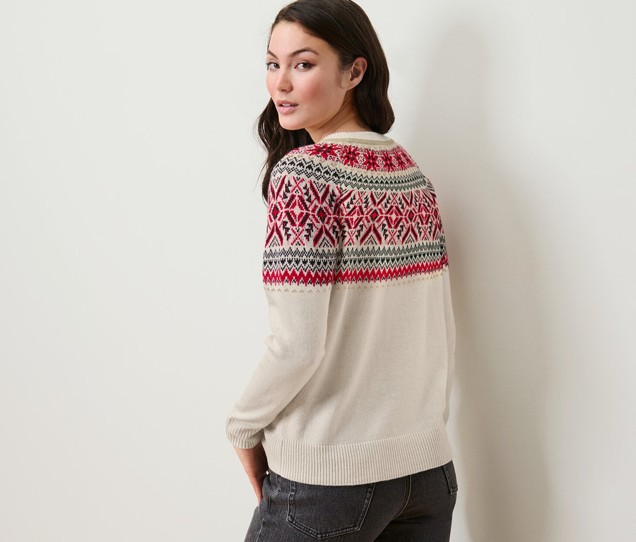 Egy nő a válla fölött hátra néz, norvég mintás kötött pulóvert és farmert visel.