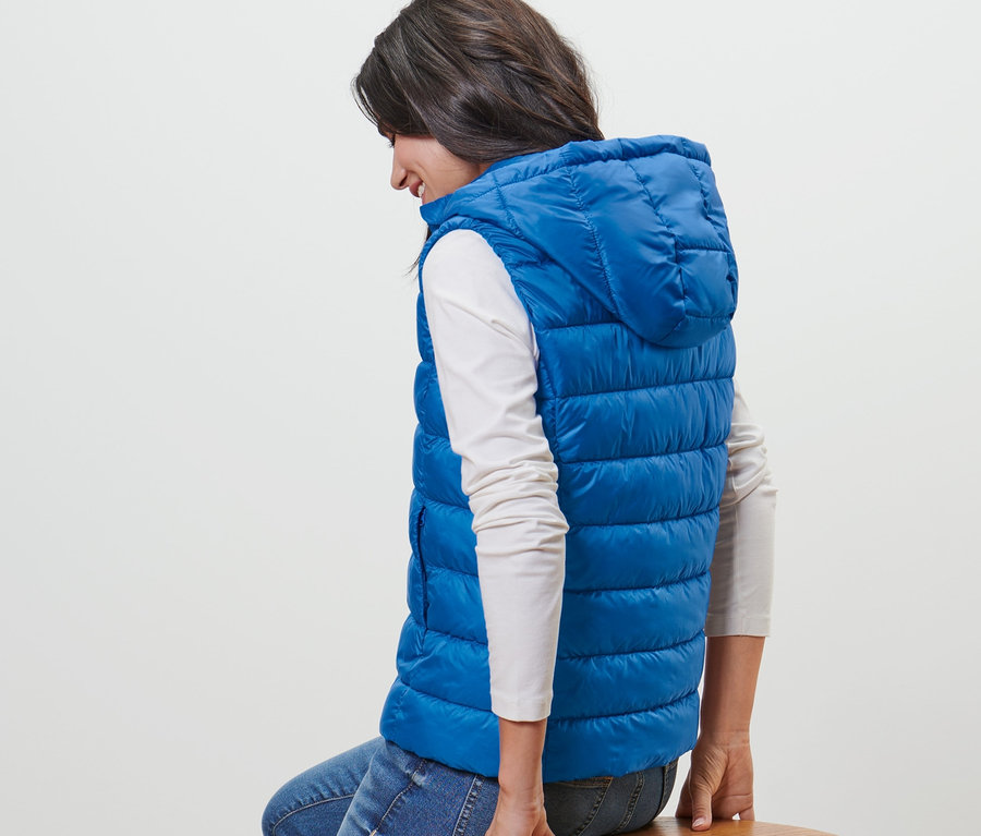 Nő ül egy fa ülőkén, kék kapucnis steppelt mellényt visel.