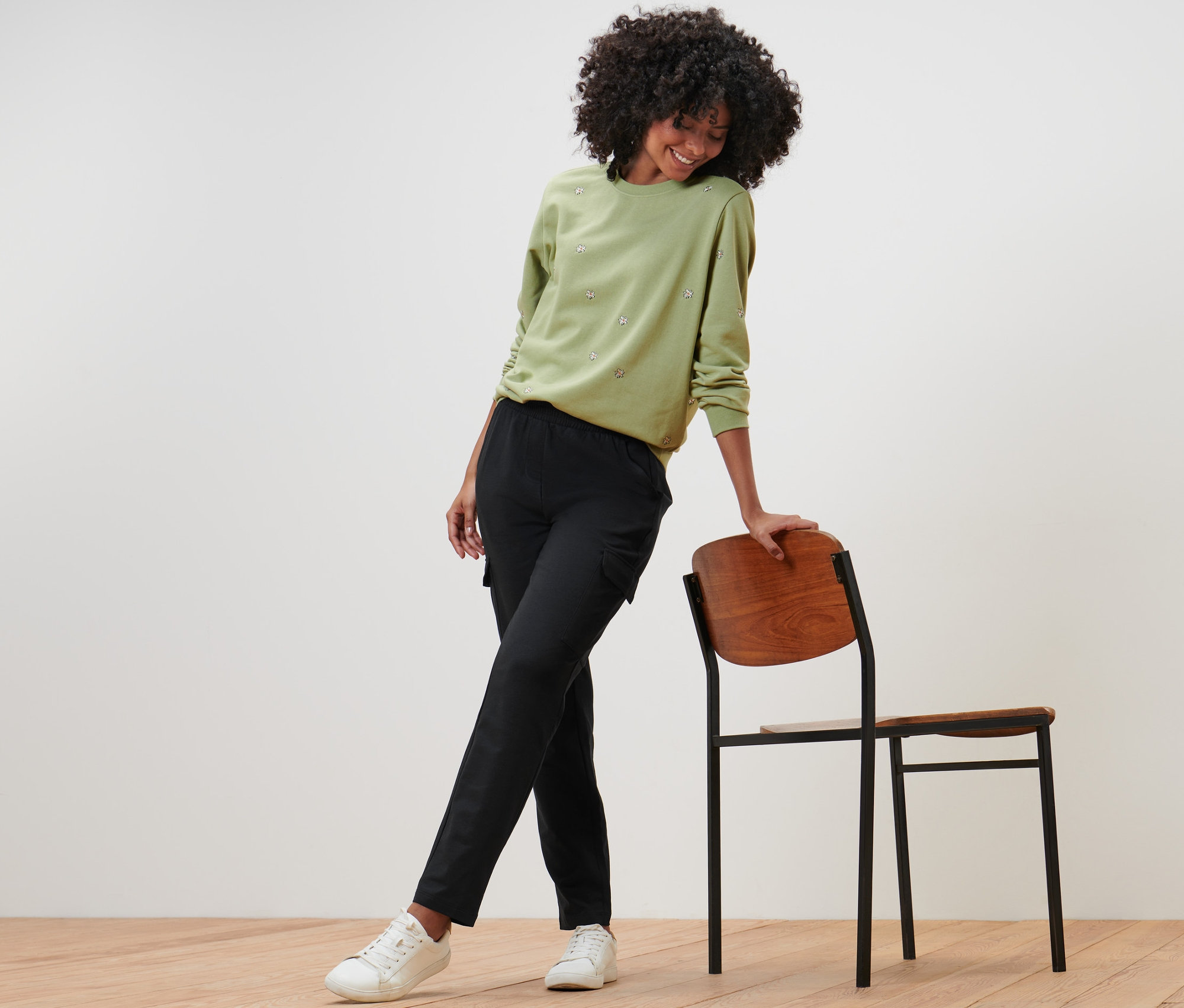 Nő támaszkodik egy széknek, zöld pulóvert és fekete oldalzsebes lezser nadrágot visel.