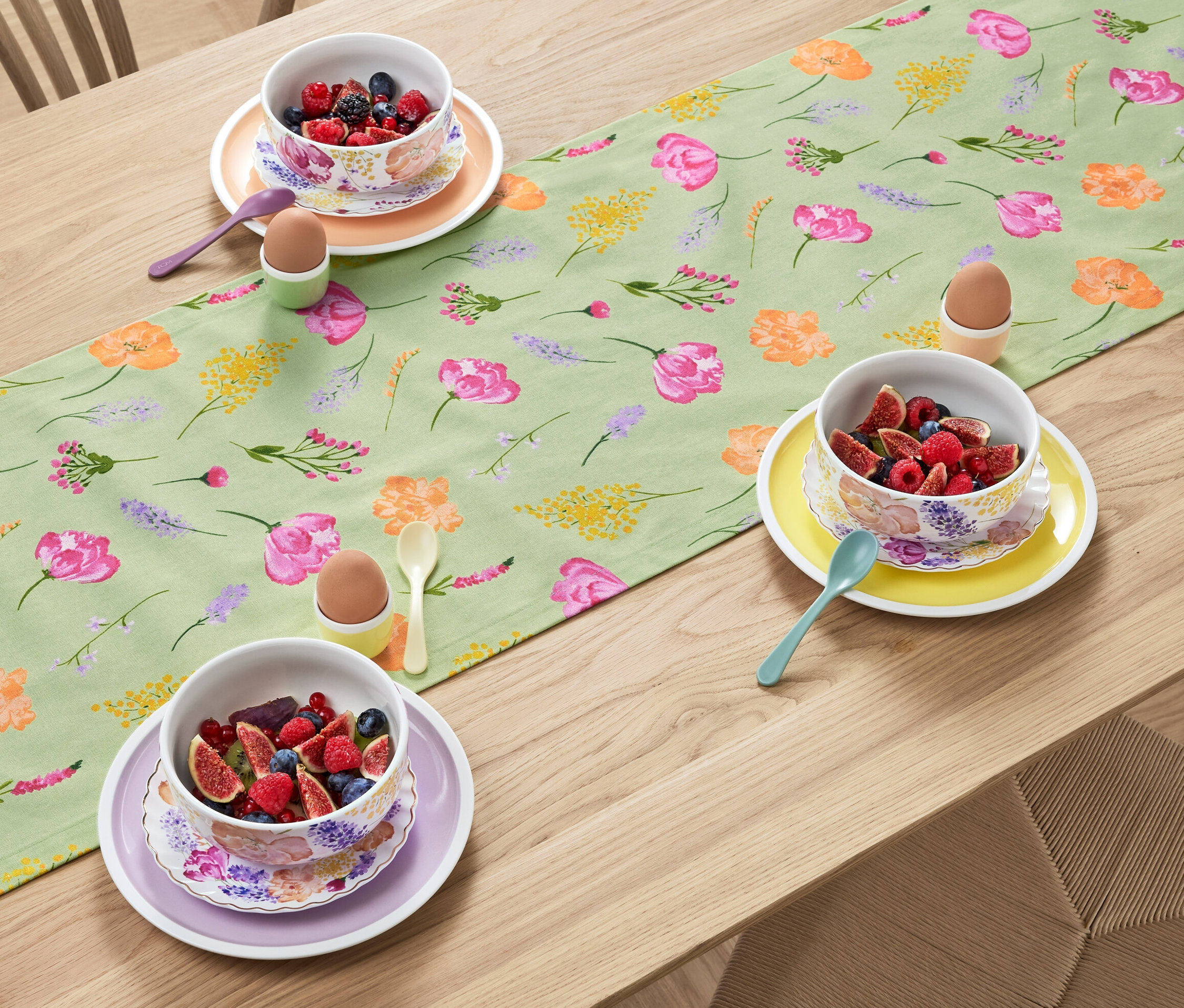 A fa asztalon egy virágmintás, zsályaszínű asztali futó található, rajta gyümölcsös tálak, tojások tartókban és kanalak.