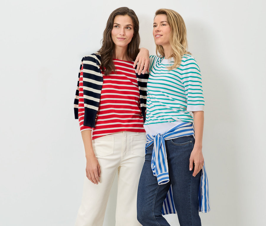 Két nő pózol fehér háttér előtt. Az egyikük víz kék finom kötésű csíkos pulóvert és farmert visel, a másikuk piros-fehér csíkos pulóvert és fehér nadrágot.