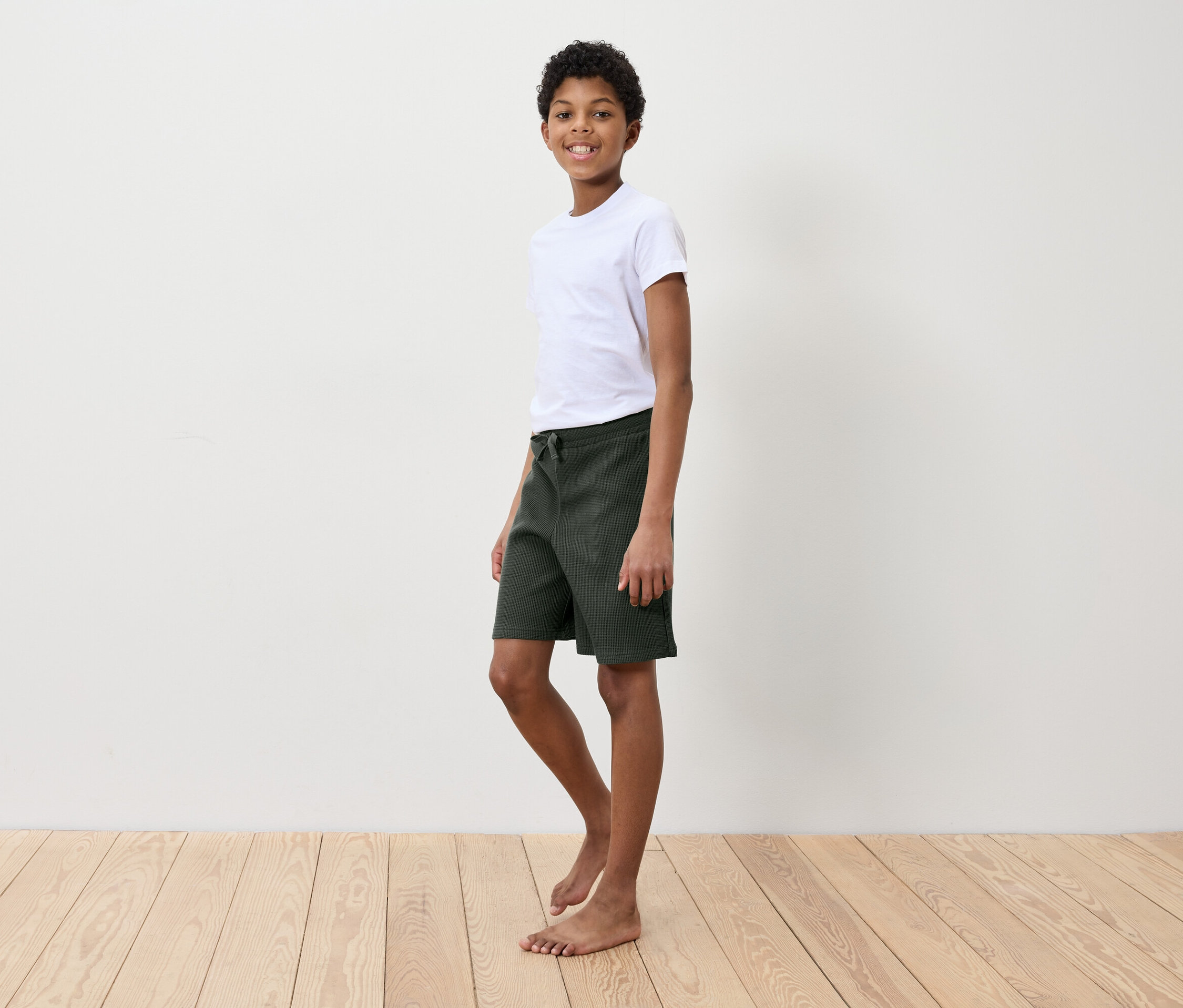 Egy fiú fehér pólóban és zöld rövidnadrágban áll egy fa padlón egy fehér fal előtt.