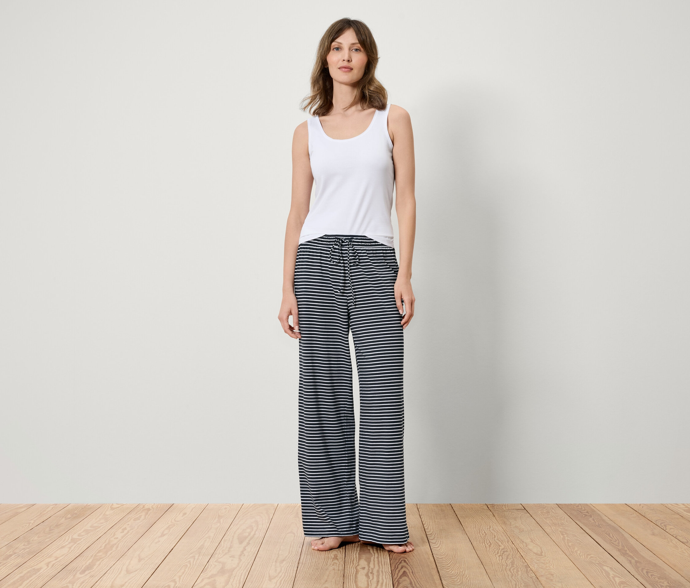 Egy nő áll egy fa padlón, fehér trikót és kék-fehér csíkos nadrágot visel.