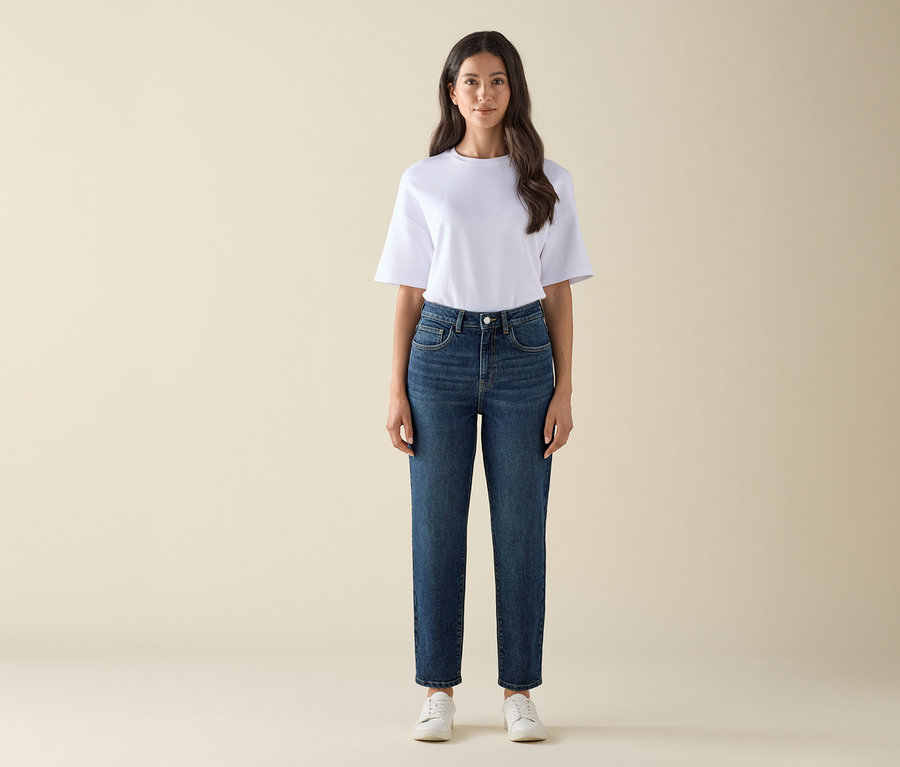 Nő áll és sötétkék »Nyla« típusú mom farmert, fehér pólót és fehér tornacipőt visel.