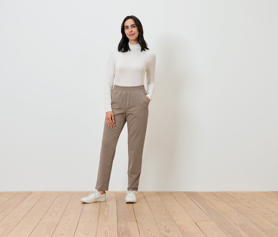 Egy nő krém színű garbót és barna nadrágot visel, miközben pózol.