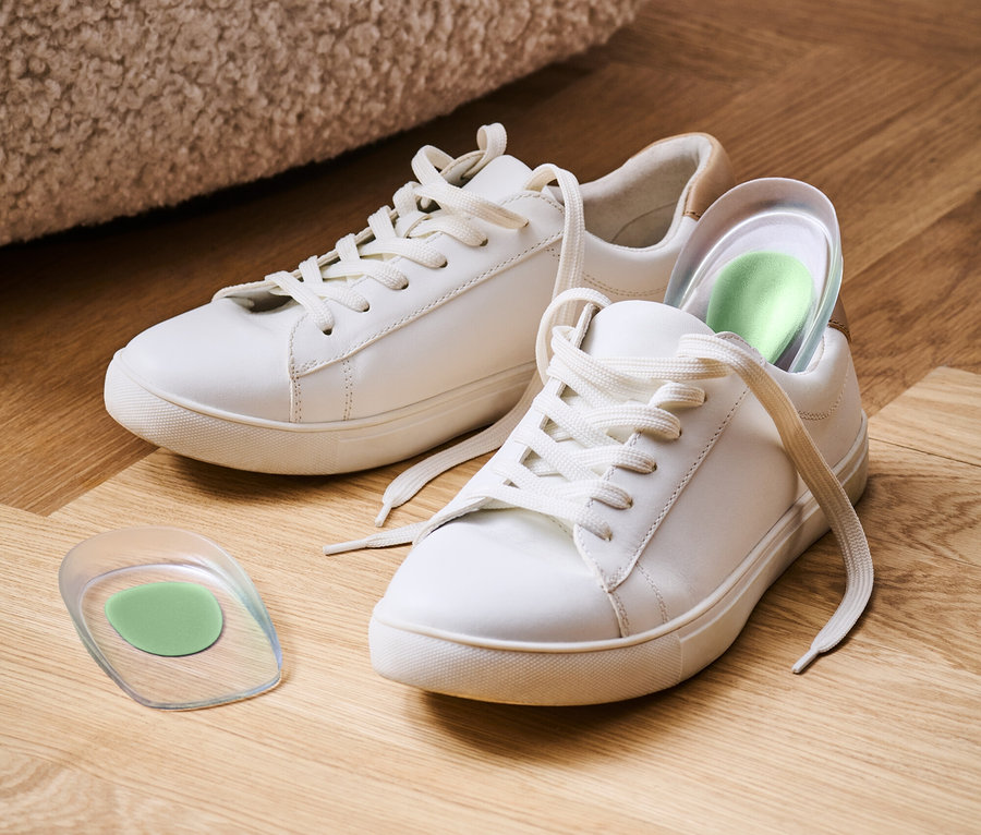 Két fehér tornacipő fekszik egy fapadlón, az egyikben zöld talpbetét van.