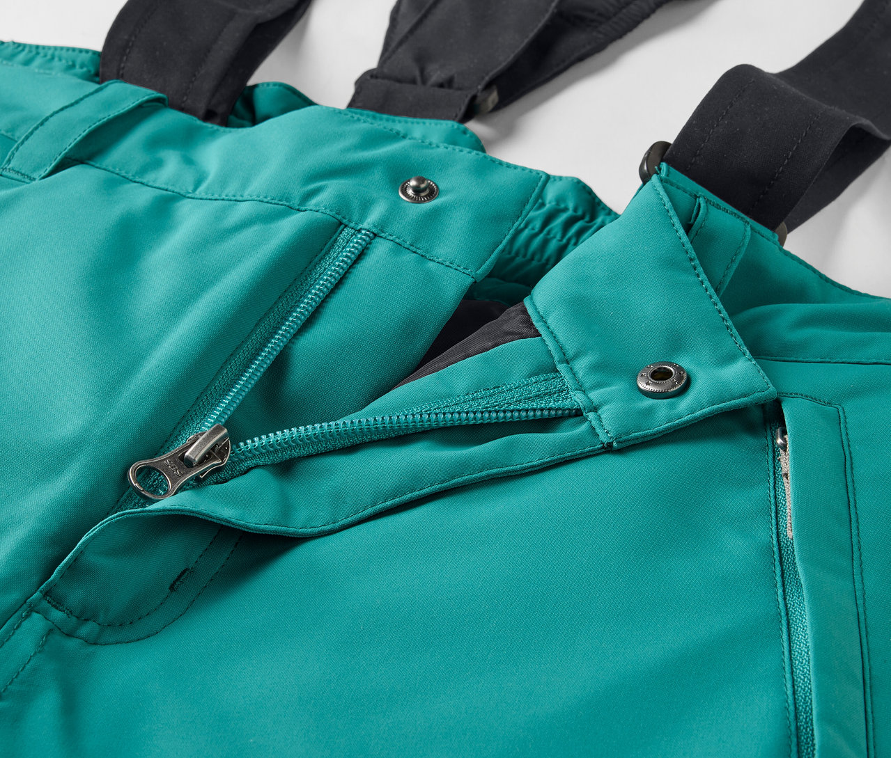 Egy zöld síkabát részlete fekete nadrágtartókkal és nyitott cipzárral.