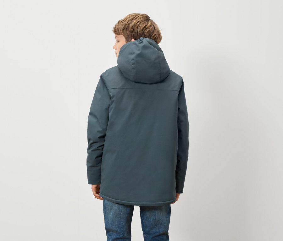 Egy kék kapucnis kabátot és kék farmert viselő fiú háttal áll a kamerának, fehér háttér előtt.