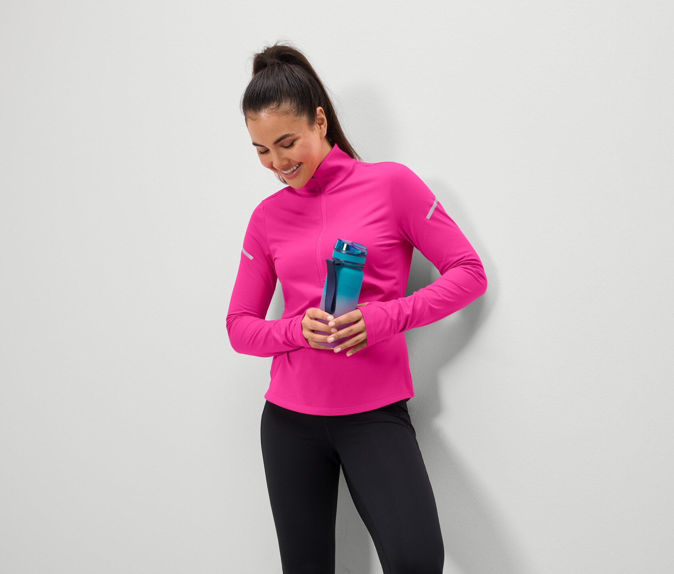 Mosolygó nő kék-zöld vizespalackot tart, rózsaszín felsőt és fekete leggingst visel.