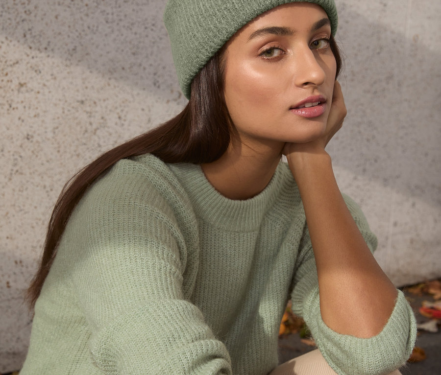 Nő ül, és zsályazöld, laza kötésű pulóvert és hozzáillő sapkát visel.
