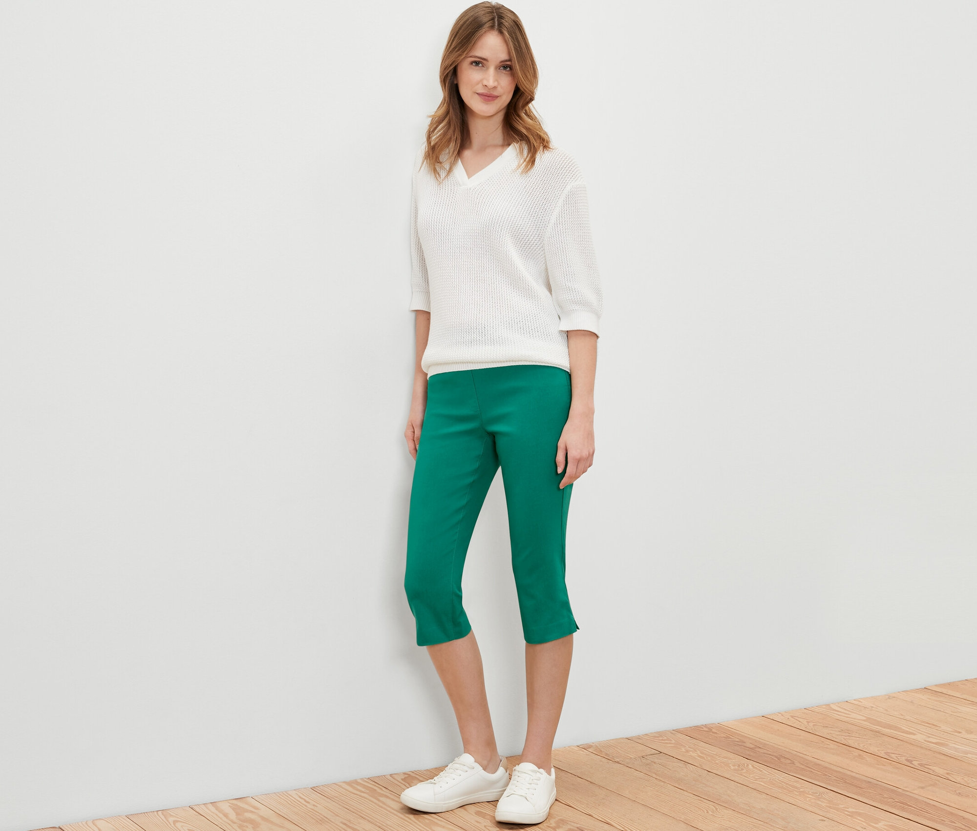Nő pózol zöld 3/4-es bengalin nadrágban, fehér pulóverben és fehér cipőben.
