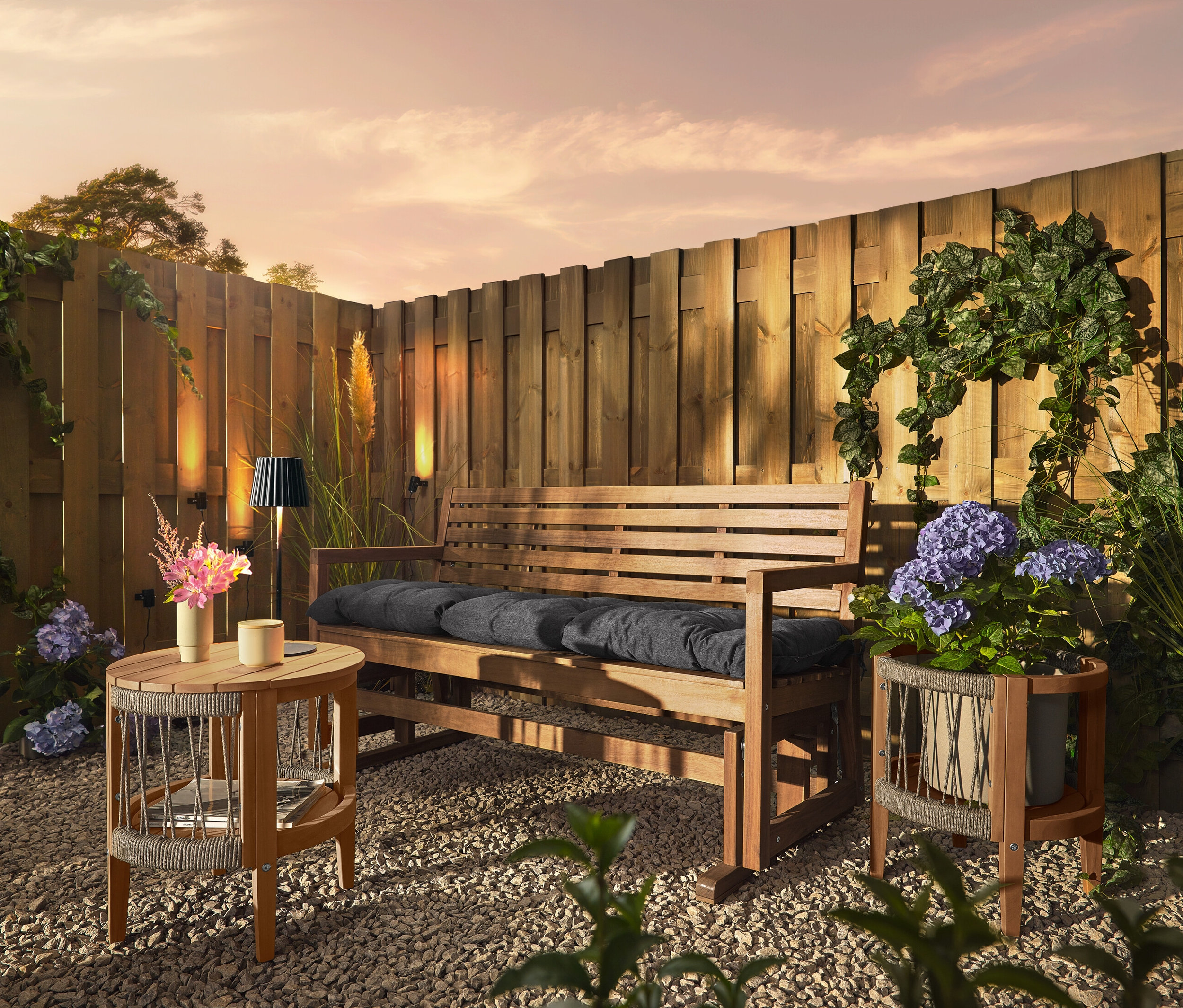 Egy fa pad párnákkal egy kavicsos talajon áll egy fa kerítés előtt. A kerítésen 3 napelemes ereszcsatorna-lámpa van rögzítve.