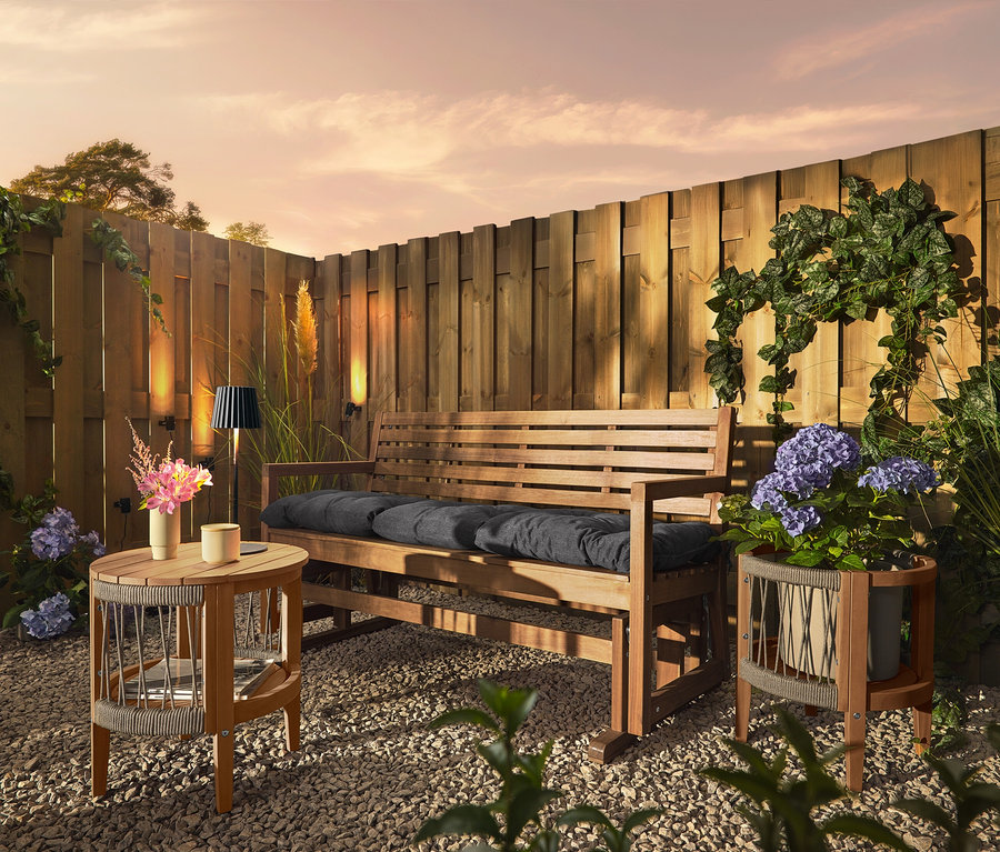 Egy fa pad párnákkal egy kavicsos talajon áll egy fa kerítés előtt. A kerítésen 3 napelemes ereszcsatorna-lámpa van rögzítve.