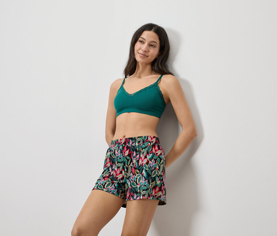 Nő a falnak dőlve, zöld csipkés bralette-et és virágmintás rövidnadrágot visel.