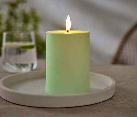 Egy zöld gyertya áll egy fehér tányéron. A háttérben egy pohár víz látható lime-mal.
