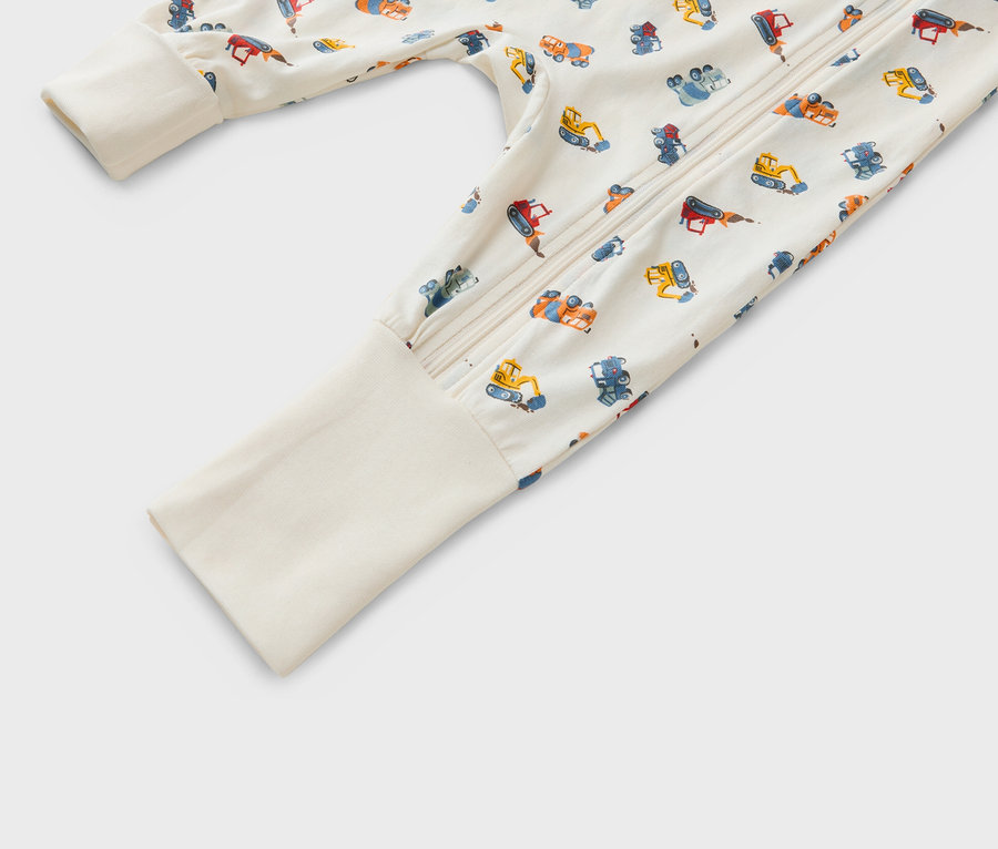 Egy bézs alapon különböző építőipari járművek mintázatával ellátott, cipzáras babapizsama közeli képe.