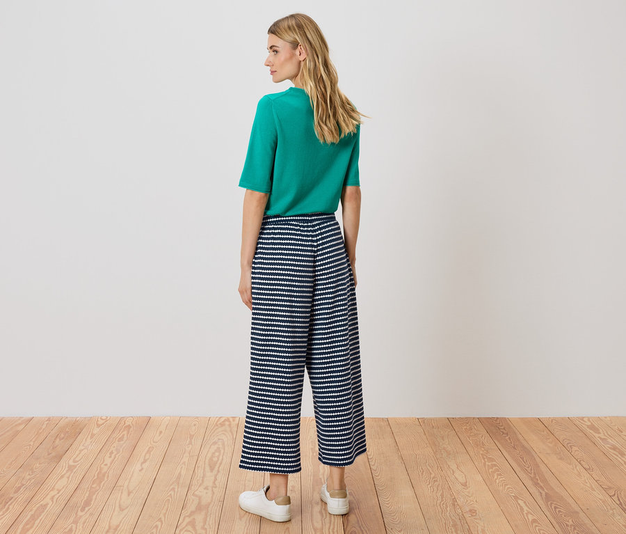 Egy nő áll háttal a kamerának, zöld felsőt és kék-fehér csíkos culottes nadrágot visel.