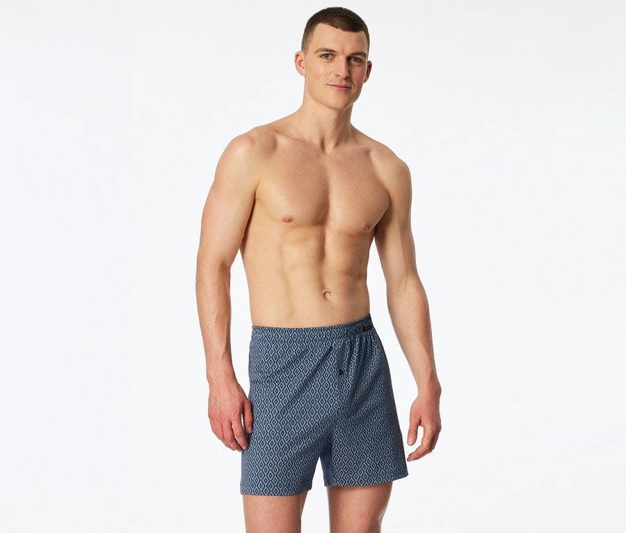 Egy félmeztelen férfi kék, rombuszmintás SCHIESSER jersey boxeralsót visel.