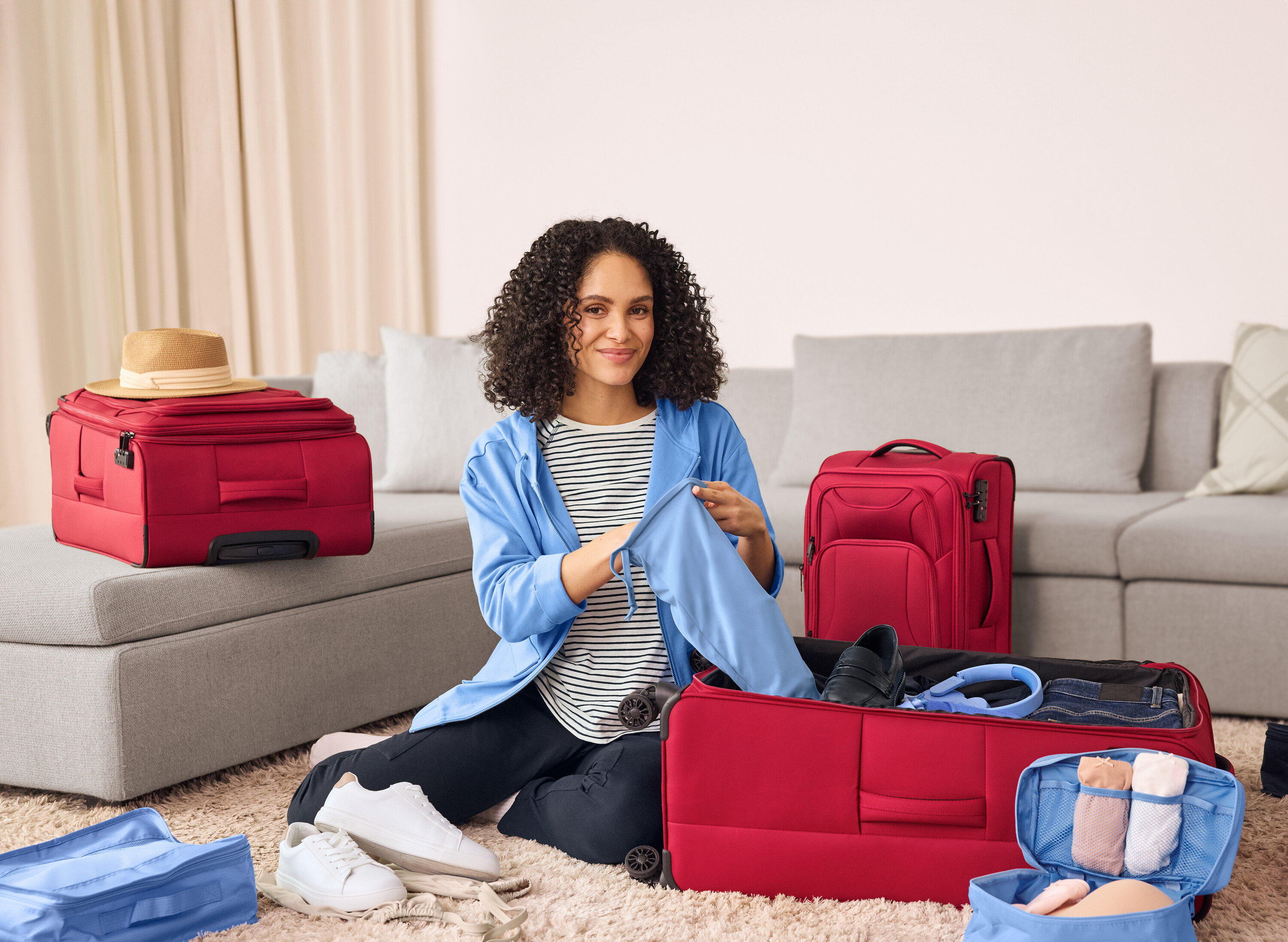 Egy nő a földön ül és sötétkék cargo zsebes joggnadrágot pakol egy piros bőröndbe. Mellette bőr sneaker cipők vannak.