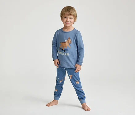 Egy mosolygó kisfiú mezítláb áll egy kék pizsamában, melyen egy gördeszkázó kutya és a "TOO COOL TO SLEEP" felirat látható.