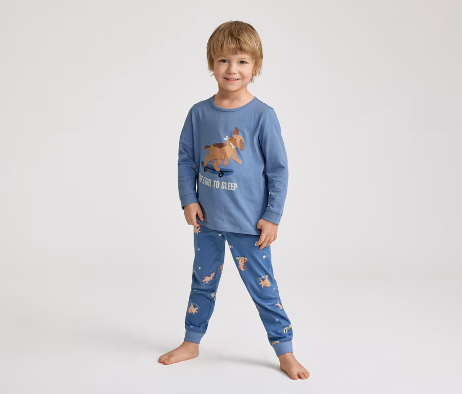 Egy mosolygó kisfiú mezítláb áll egy kék pizsamában, melyen egy gördeszkázó kutya és a "TOO COOL TO SLEEP" felirat látható.