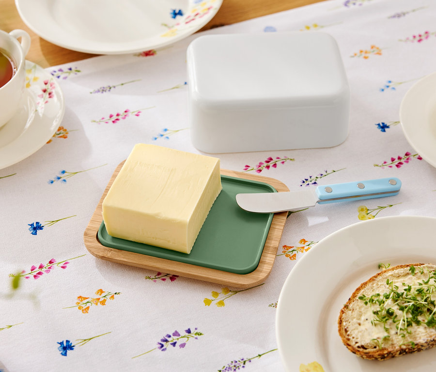 A virágos asztalon vaj van egy zöld betétes fa tányéron, egy vajkés, egy vajas doboz, egy csésze tea és egy tányér egy vajas szendviccsel.