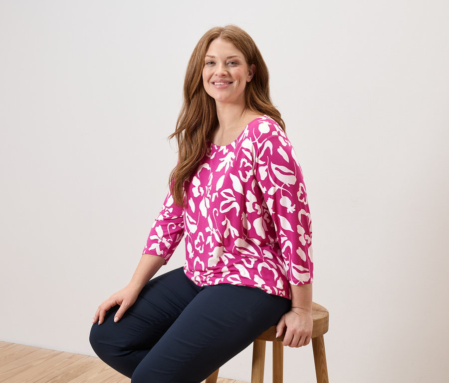 Egy hosszú, vörös hajú nő ül egy fa széken, rózsaszín, fehér leveles blúzt és sötétkék nadrágot visel.