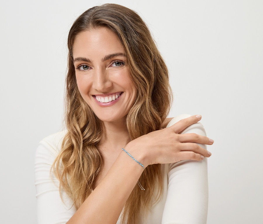 Mosolygó nő szőke, hullámos hajjal és kék karkötővel a bal csuklóján, fehér háttér előtt.