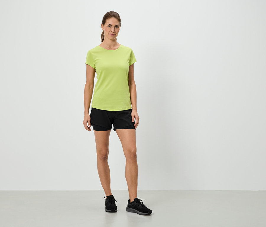 Egy nő áll egy világoszöld pólóban, fekete rövidnadrágban és fekete tornacipőben egy fehér fal előtt.