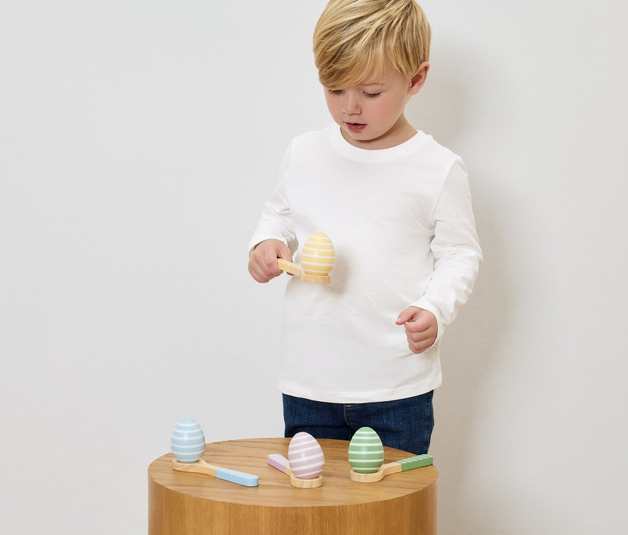 Egy fiú festett fa tojásokkal és kis kalapácsokkal játszik egy fa asztalon.