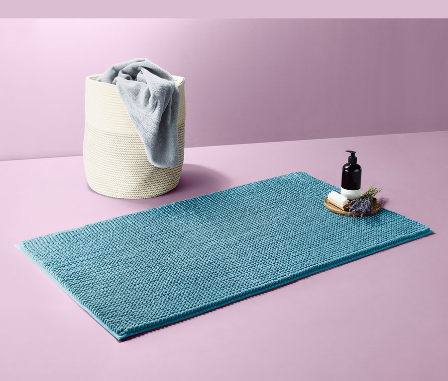 Egy kb. 70 x 120 cm-es kék kádkilépő a padlón. Mellette egy szappanadagoló, szappan és levendula található. A háttérben egy törölközőket tartalmazó kosár látható.