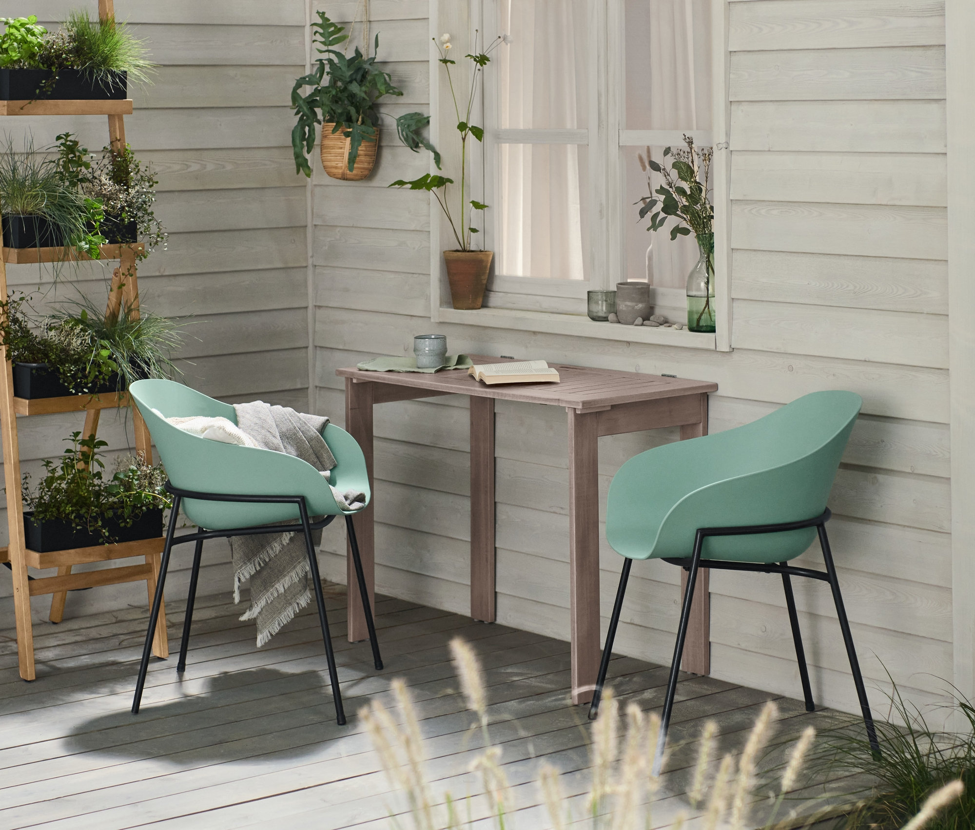 Egy teraszon egy összecsukható balkon asztal »Lenja« és két mentazöld modern kagylószék látható. A háttérben egy fekete, összecsukható növénytartó állvány ültetőládákkal.