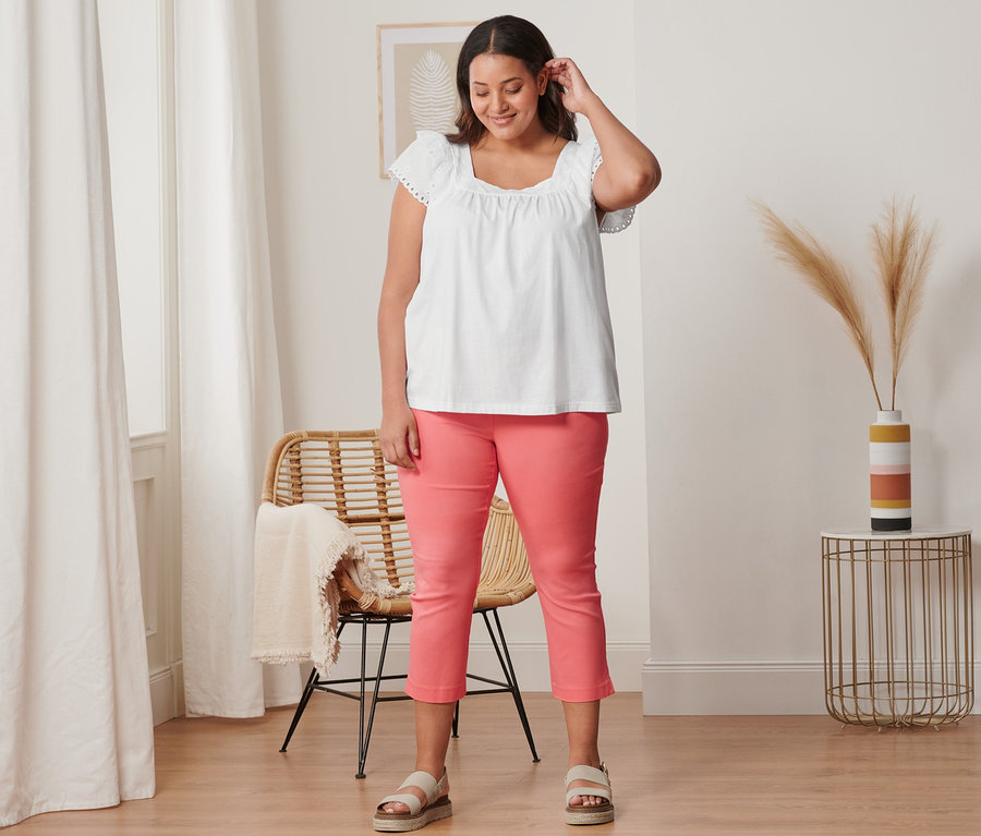 Nő visel korall színű 7/8-os sztreccsnadrágot, fehér hímzett pólót és bőr szandált.
