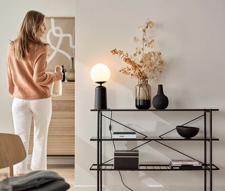 Nő egy üveget tart a kezében egy kb. 120 cm széles konzolasztal mellett, melyen egy fekete, üveggömbös asztali lámpa áll.