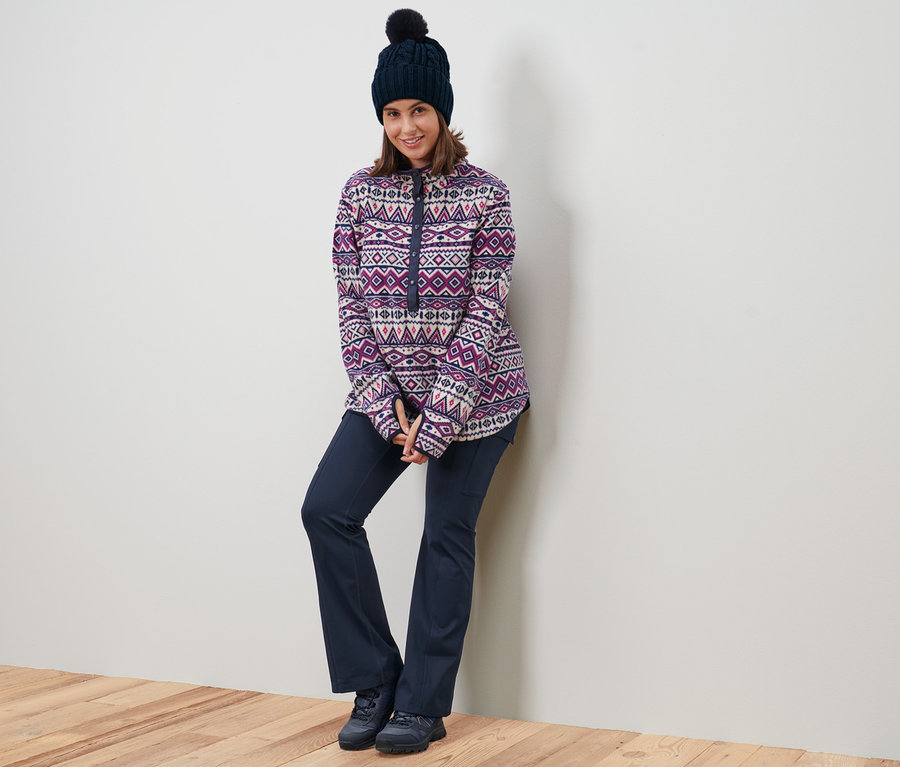 Egy nő a falnak dőlve női mikropolár pulóvert, sötétkék, polárbéléses kötött sapkát és sötétkék bőszárú termonadrágot visel.