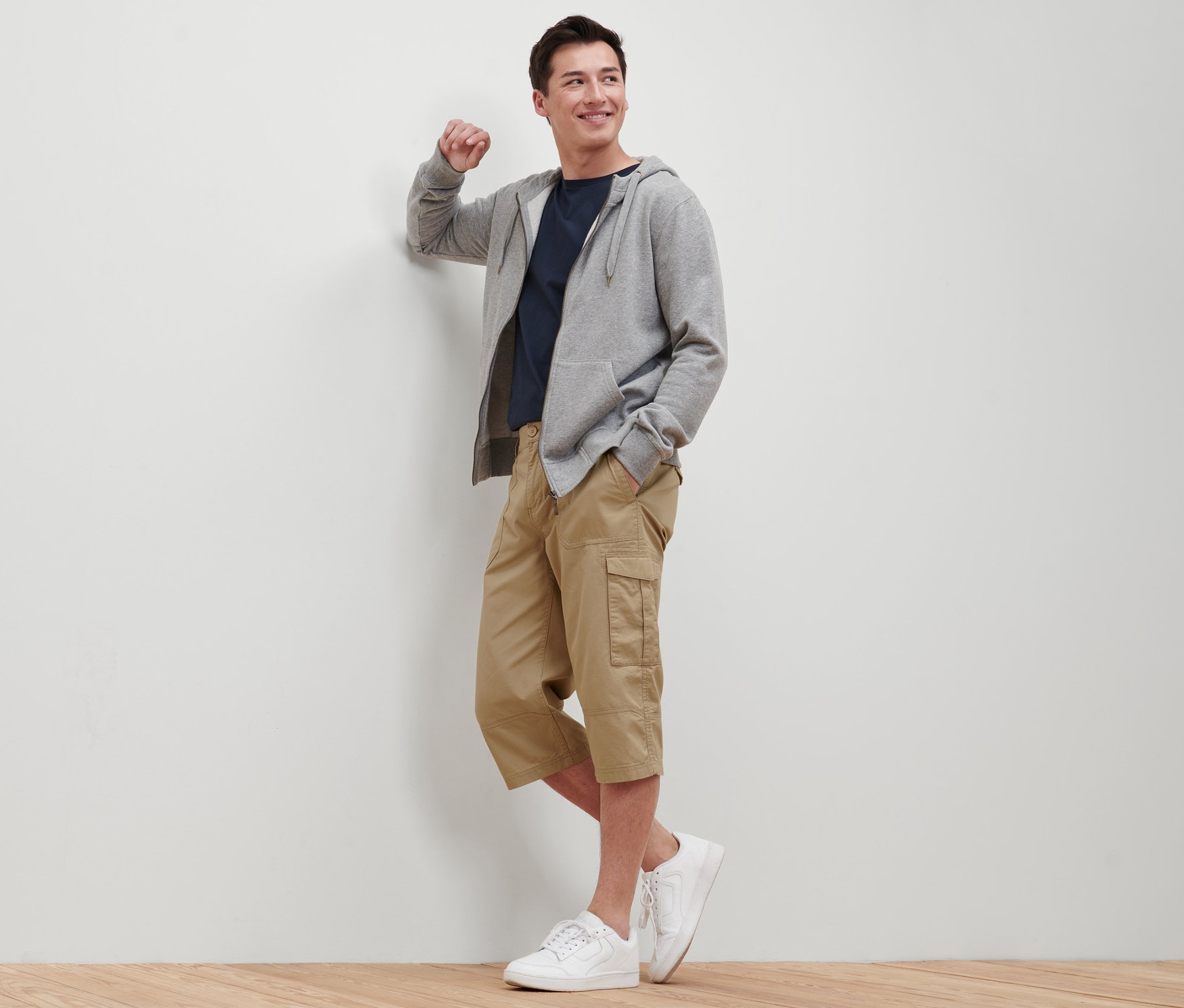 Férfi a falnak támaszkodva, bézs színű, 3/4-es cargo bermudanadrágot, pólót, kapucnis pulóvert és tornacipőt visel.