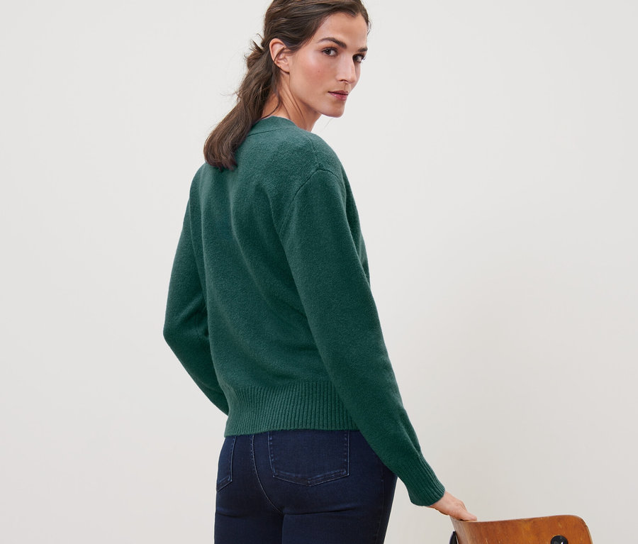 Egy nő a válla fölött hátra néz, sötétzöld, gyapjú kötött kardigánt és farmert visel.