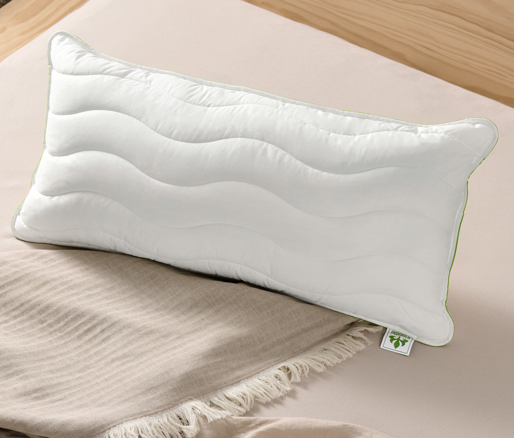 Egy fehér irisette® greenline Megafil® BIO párna, kb. 40 x 80 cm méretben egy bézs színű ágytakarón fekszik egy ágyon.