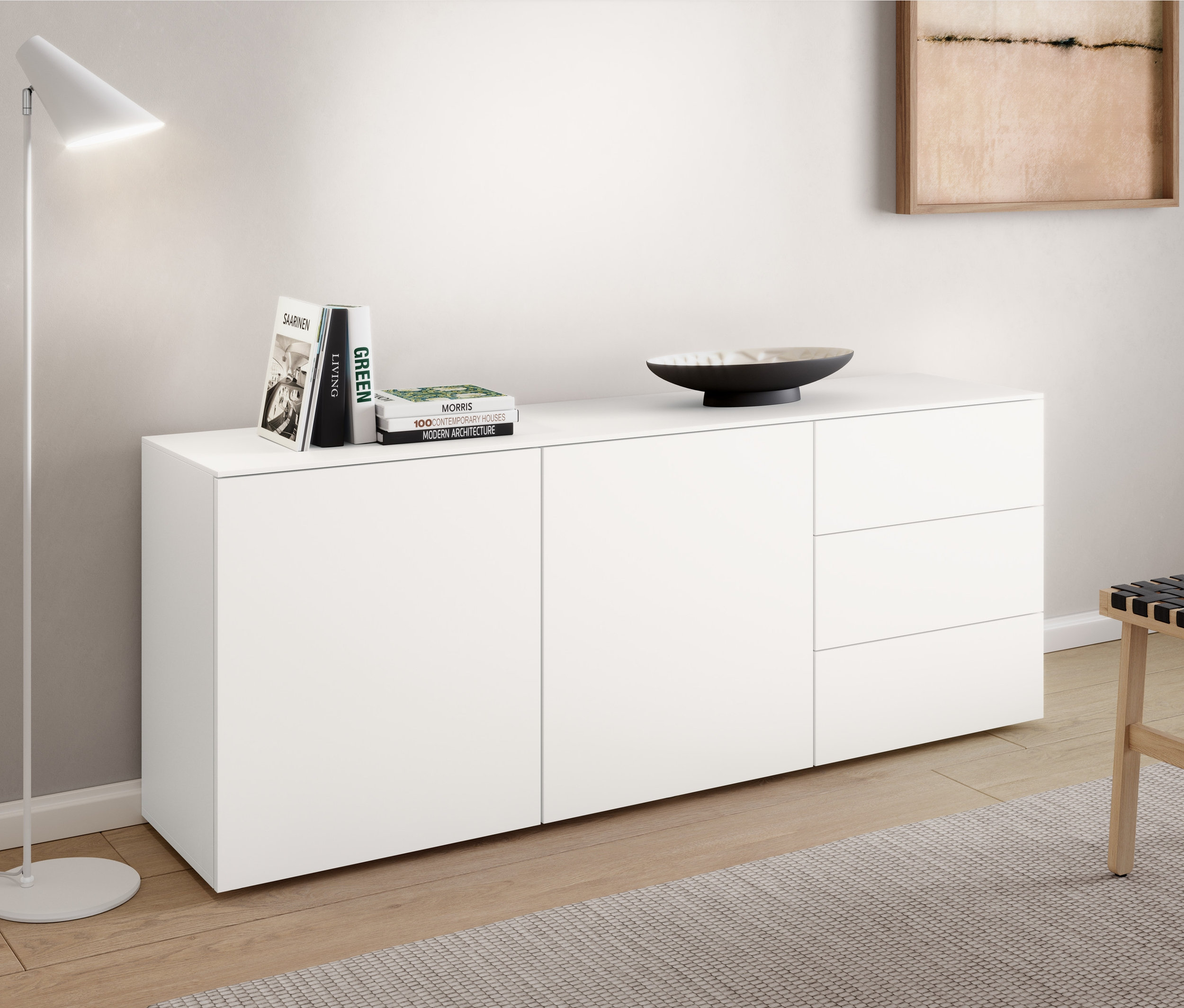 Egy fehér »Cale« fém állólámpa áll egy komód mellett. A komódon könyvek és egy tál található. A komód mellett egy fonott ülőke áll.