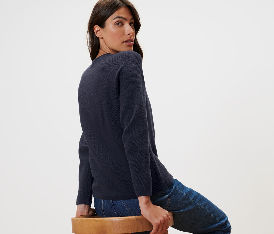 Nő ül oldalra egy fa székre néz a válla fölött. Sötétkék kötött pulóvert visel.