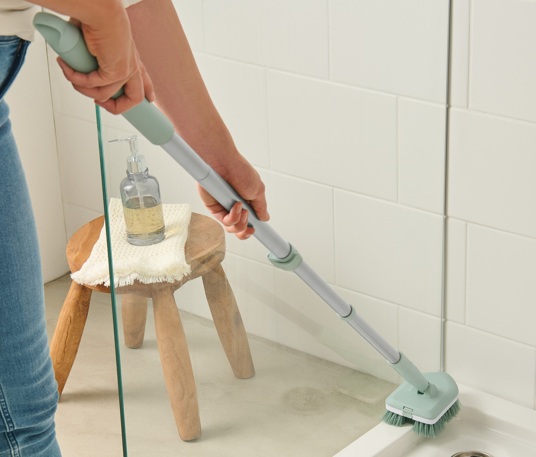 Egy nő csempetisztító kefével tisztít egy zuhanykabint. Egy sámlira szappanadagoló és törölköző van helyezve.