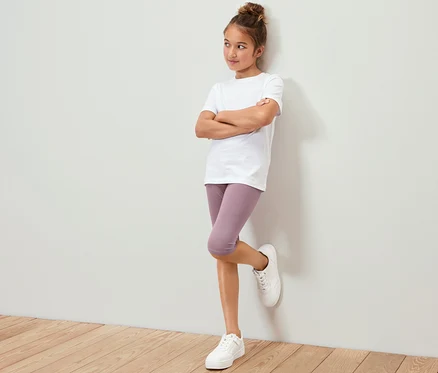 Egy lány fehér pólóban és lila 3/4-es leggingsben (3 lány 3/4-es leggings, világoskék/mályva) a falnak dőlve áll.