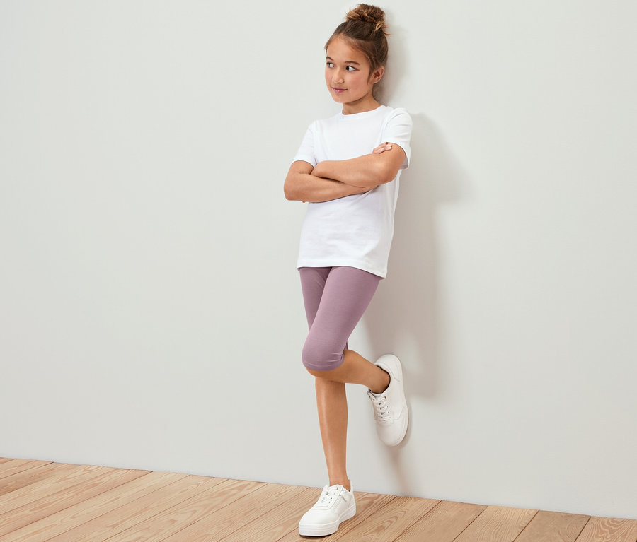 Egy lány fehér pólóban és lila 3/4-es leggingsben (3 lány 3/4-es leggings, világoskék/mályva) a falnak dőlve áll.