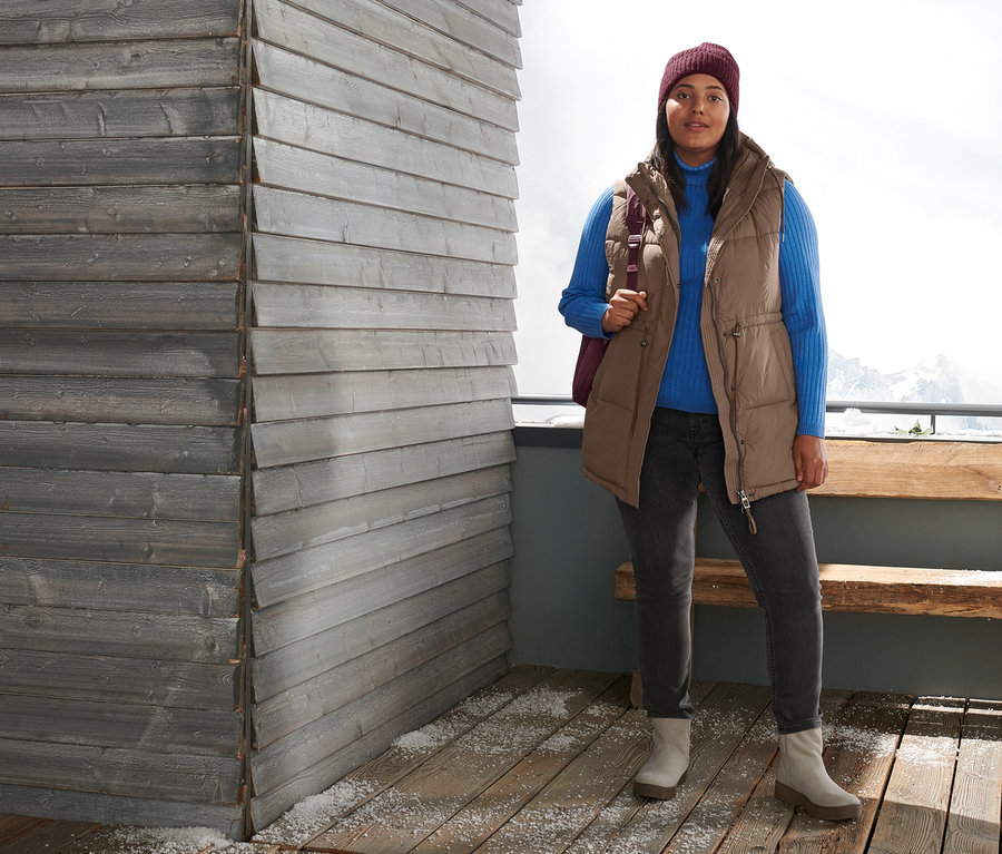 Egy nő áll kint, és steppelt mellényt, bordázott garbónyakú pulóvert, slimfit farmert, velúrbőr csizmát és bordó kötött sapkát visel. Egy steppelt hátizsákot visel.