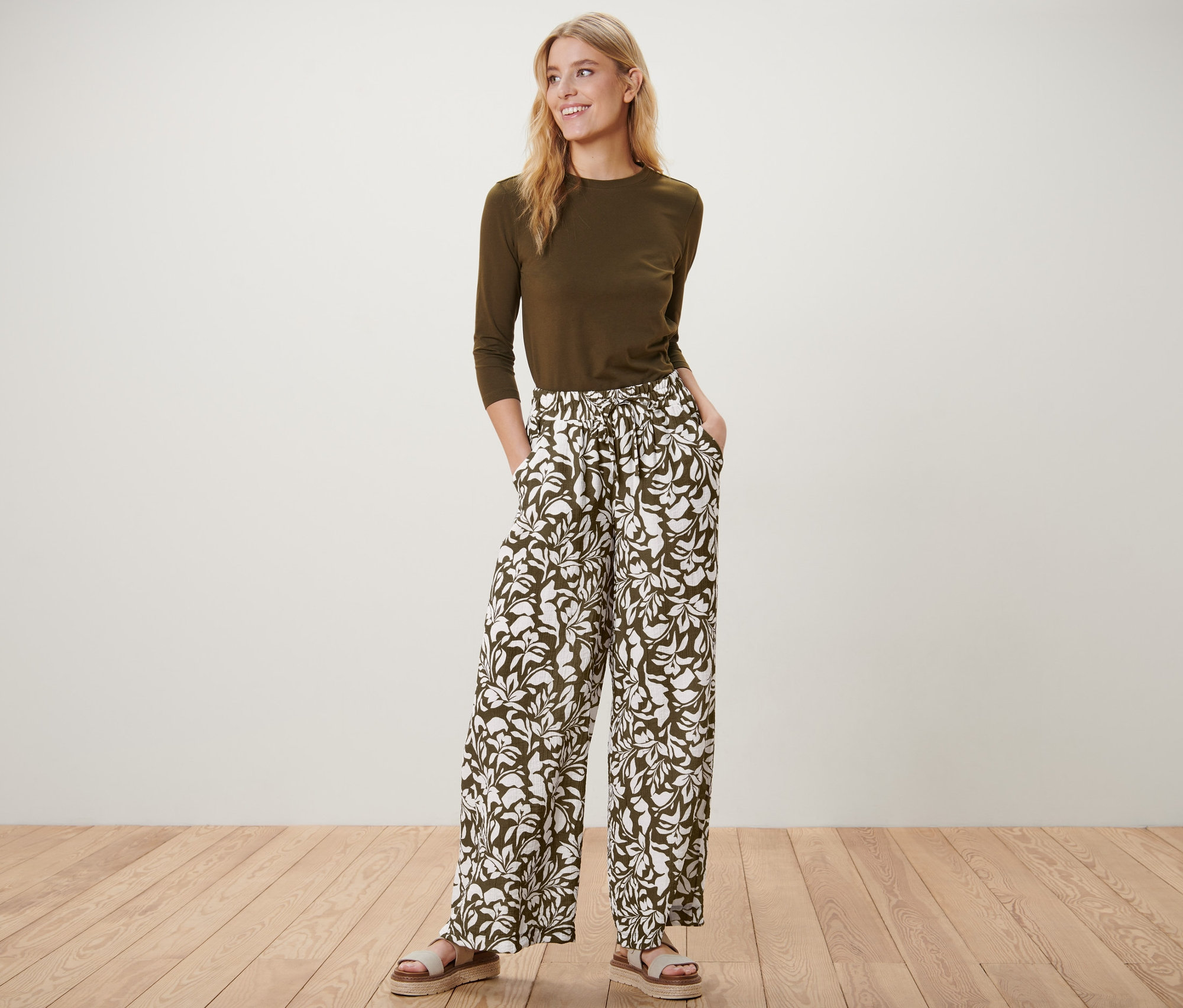 Egy nő állva pózol nyomott mintás belebújós nadrágban, olívazöld felsőben és szandálban.