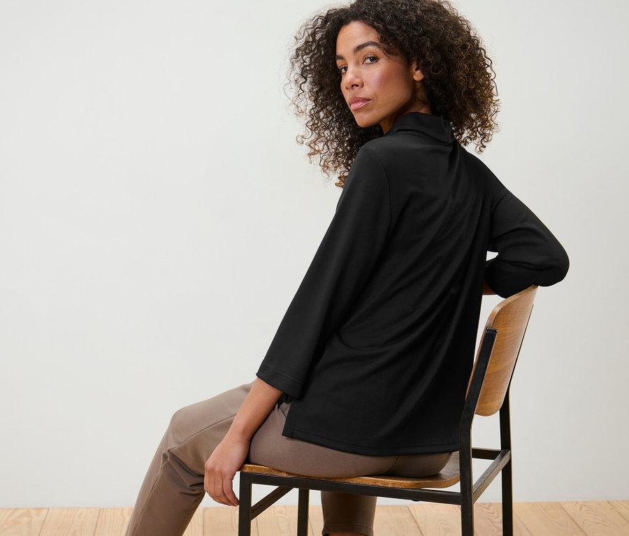 Nő ül egy széken, és fekete, 3/4-es ujjú szabadidőfelsőt visel.