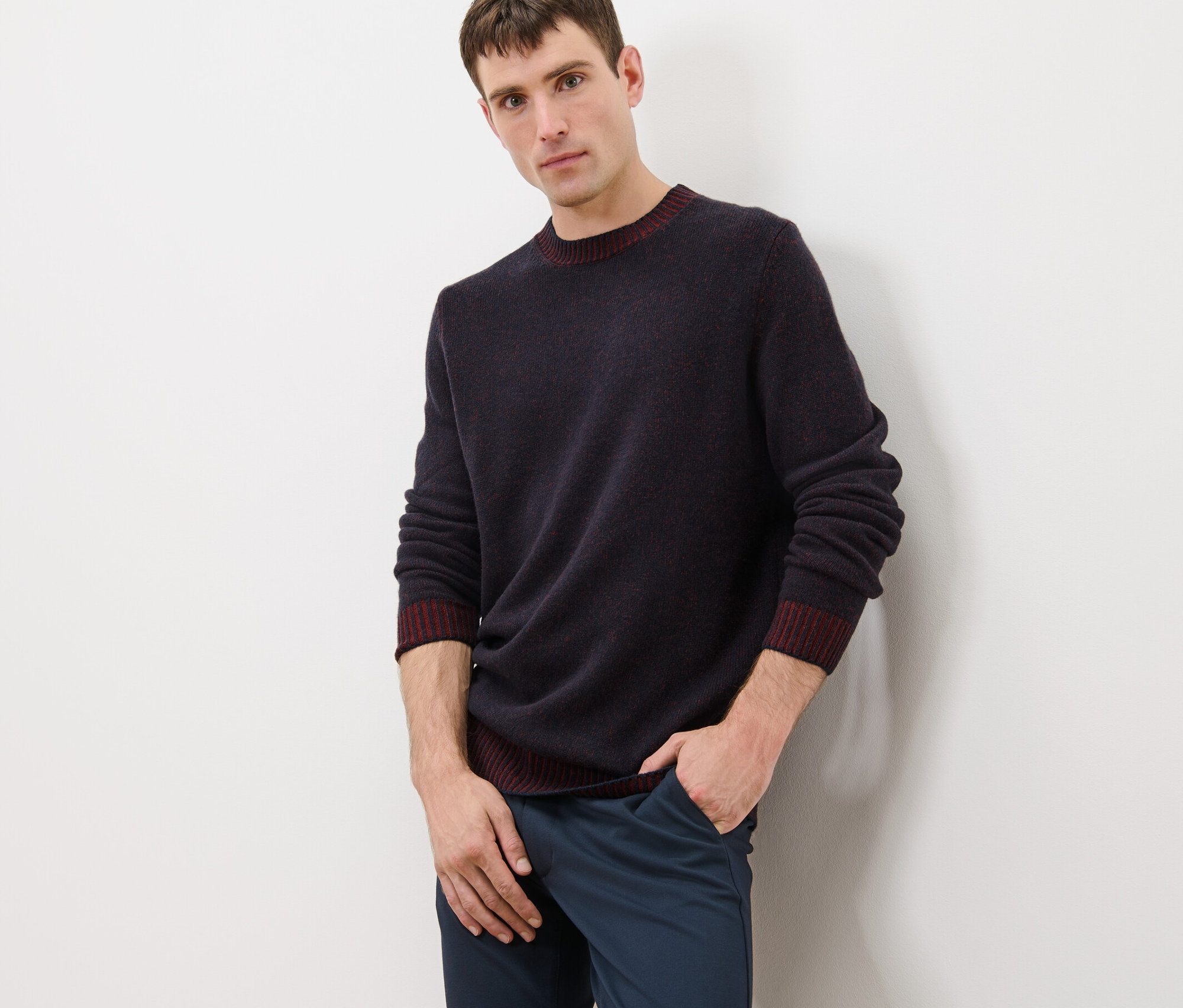 Férfi sötétkék-piros szegélyű gyapjú kötött pulóverben és kék nadrágban pózol.